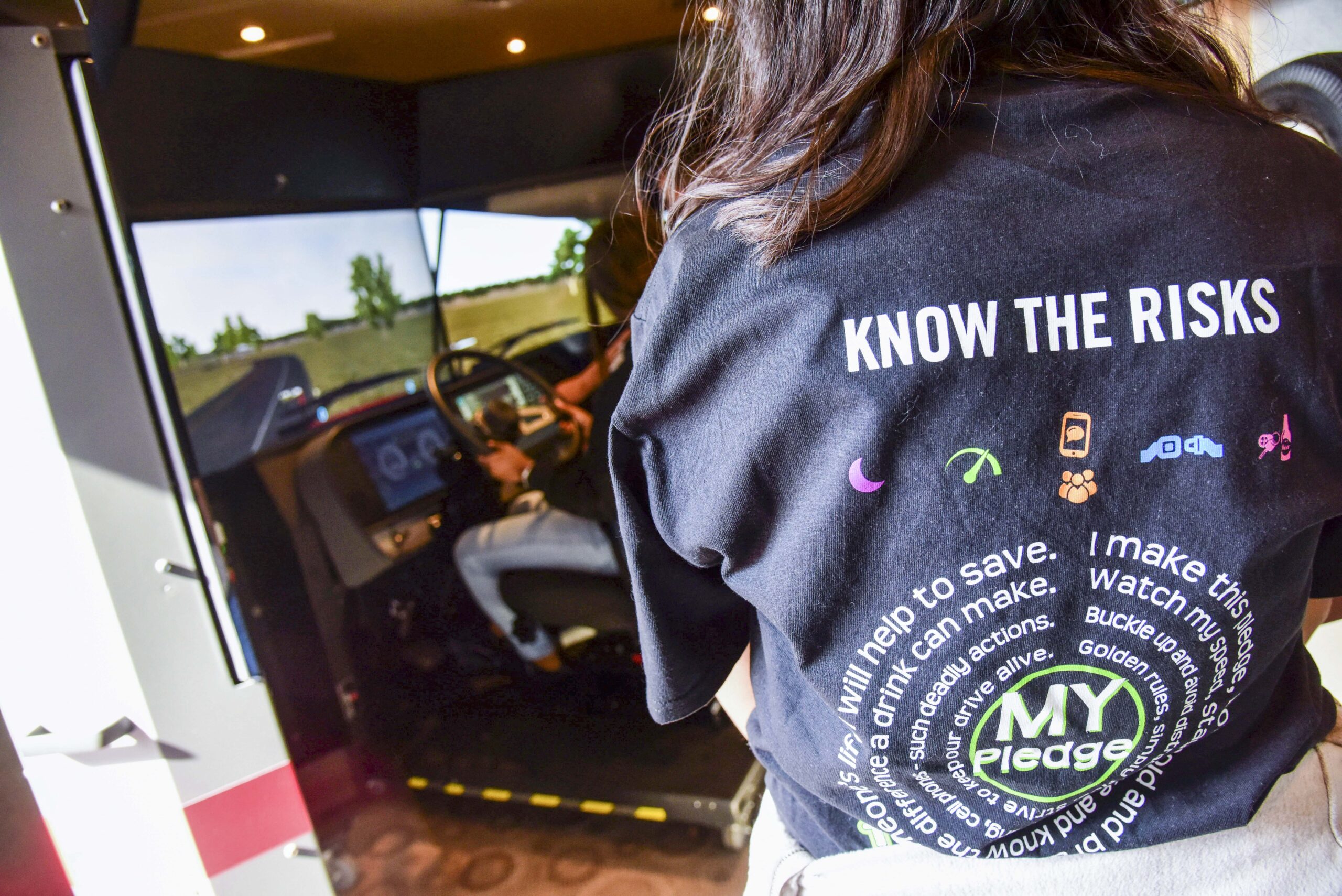 A person wearing a “Know the Risks” and “MY Pledge” shirt stands in front of another person using a driving simulator, with a road scene displayed on the simulator’s screen.