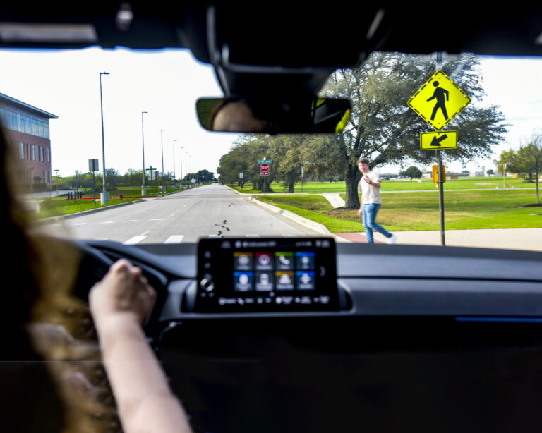 View from inside a car as a driver approaches a crosswalk with a pedestrian crossing the street and a yellow pedestrian crossing sign visible.