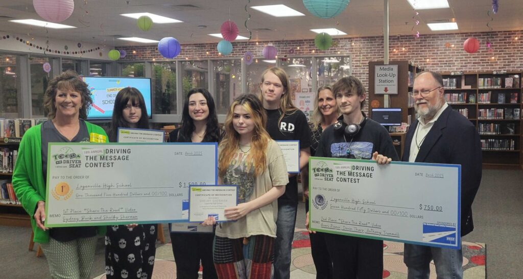 A group of teens and adults pose in a library, holding large prize checks and certificates from a driving safety contest.