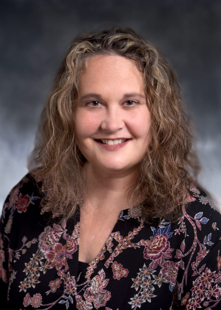 A woman with curly, shoulder-length hair and a floral blouse smiles at the camera against a neutral background.