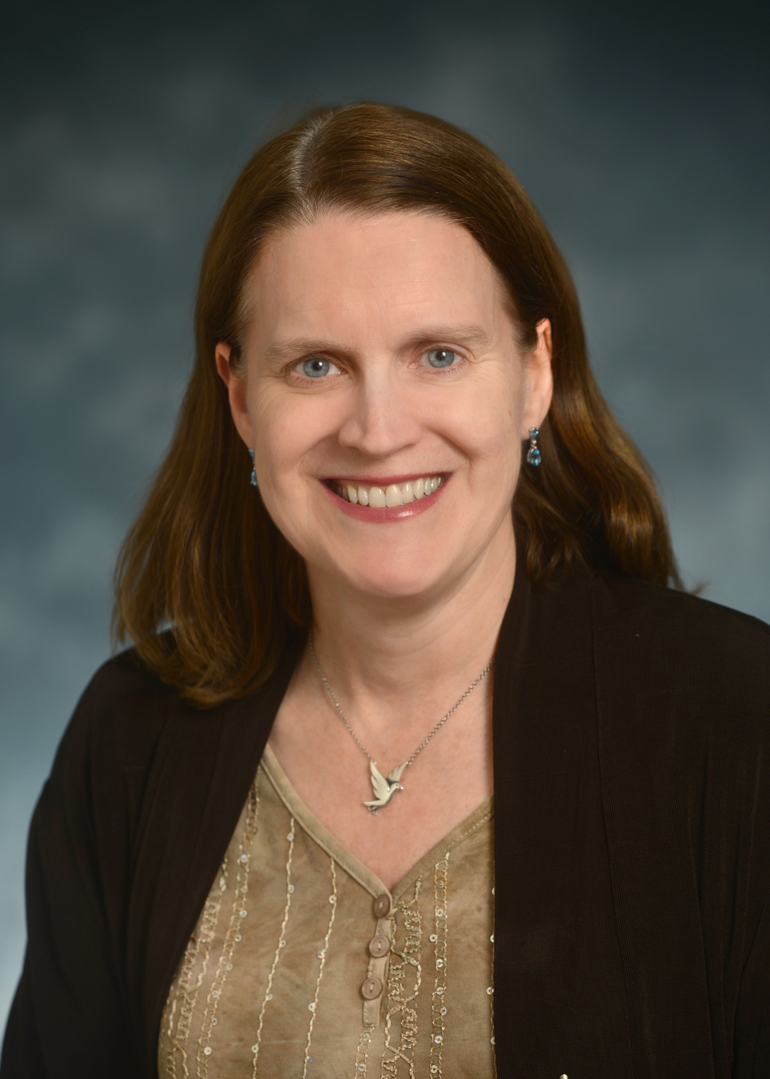 A woman with shoulder-length brown hair is smiling at the camera, wearing a brown jacket, a beige top, a bird-shaped necklace, and blue earrings, with a blurred blue-gray background.
