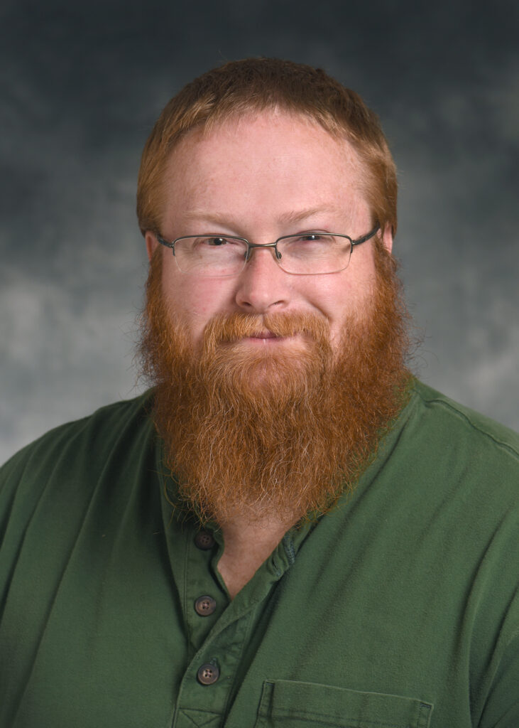 A man with a thick red beard and glasses wearing a green shirt, posed against a neutral, blurred background.