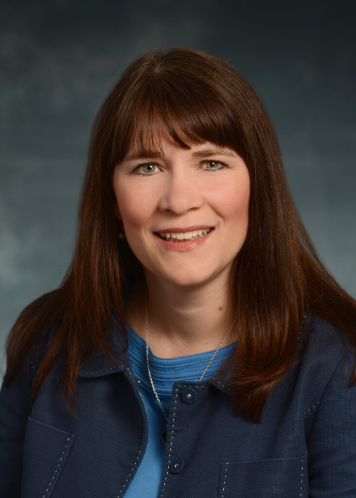 Woman with long brown hair wearing a blue top and jacket, smiling at the camera against a neutral background.