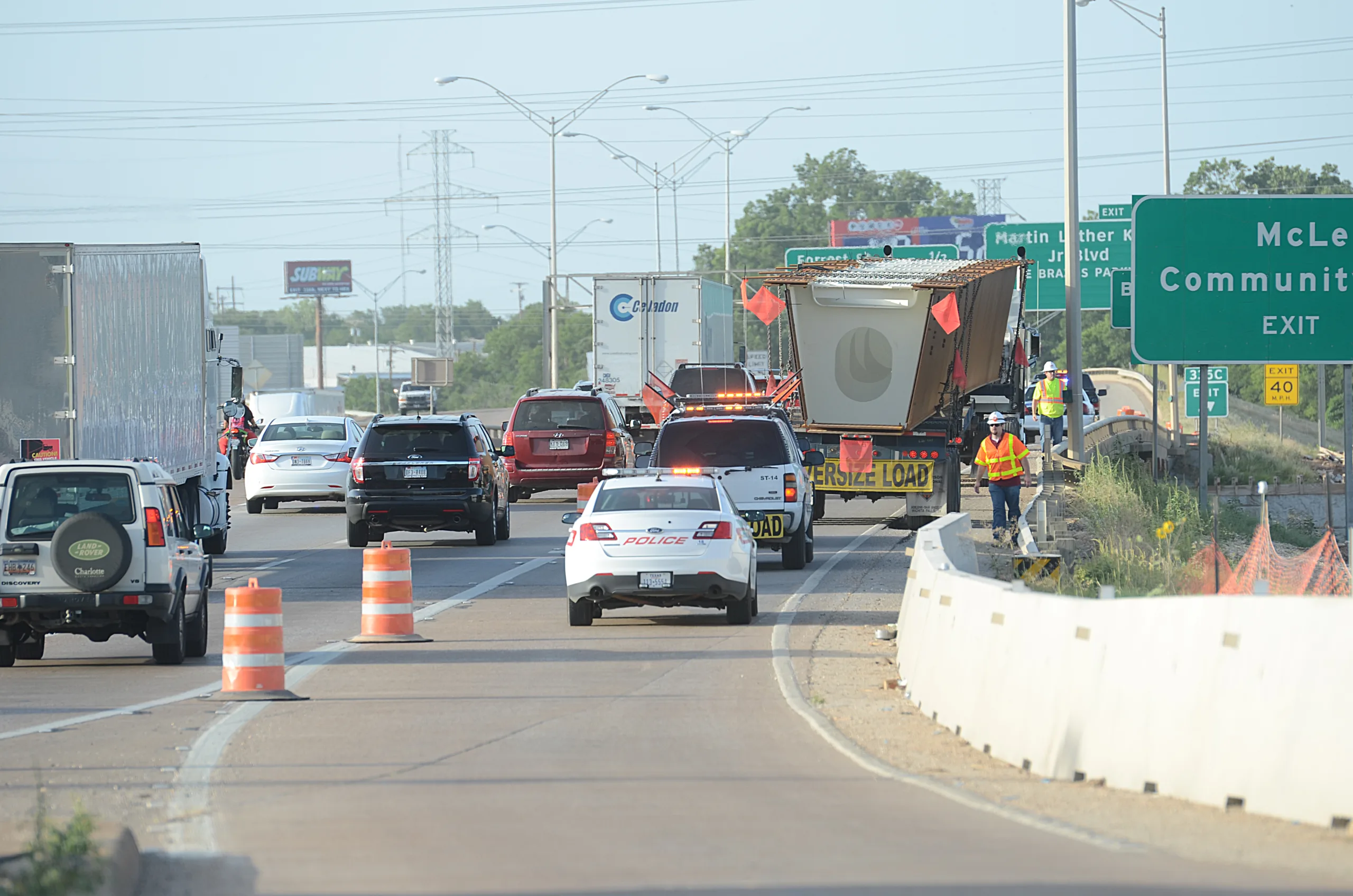 Police vehicles and traffic surround an oversized load with escort vehicles on a busy highway near an exit; construction cones line the road.