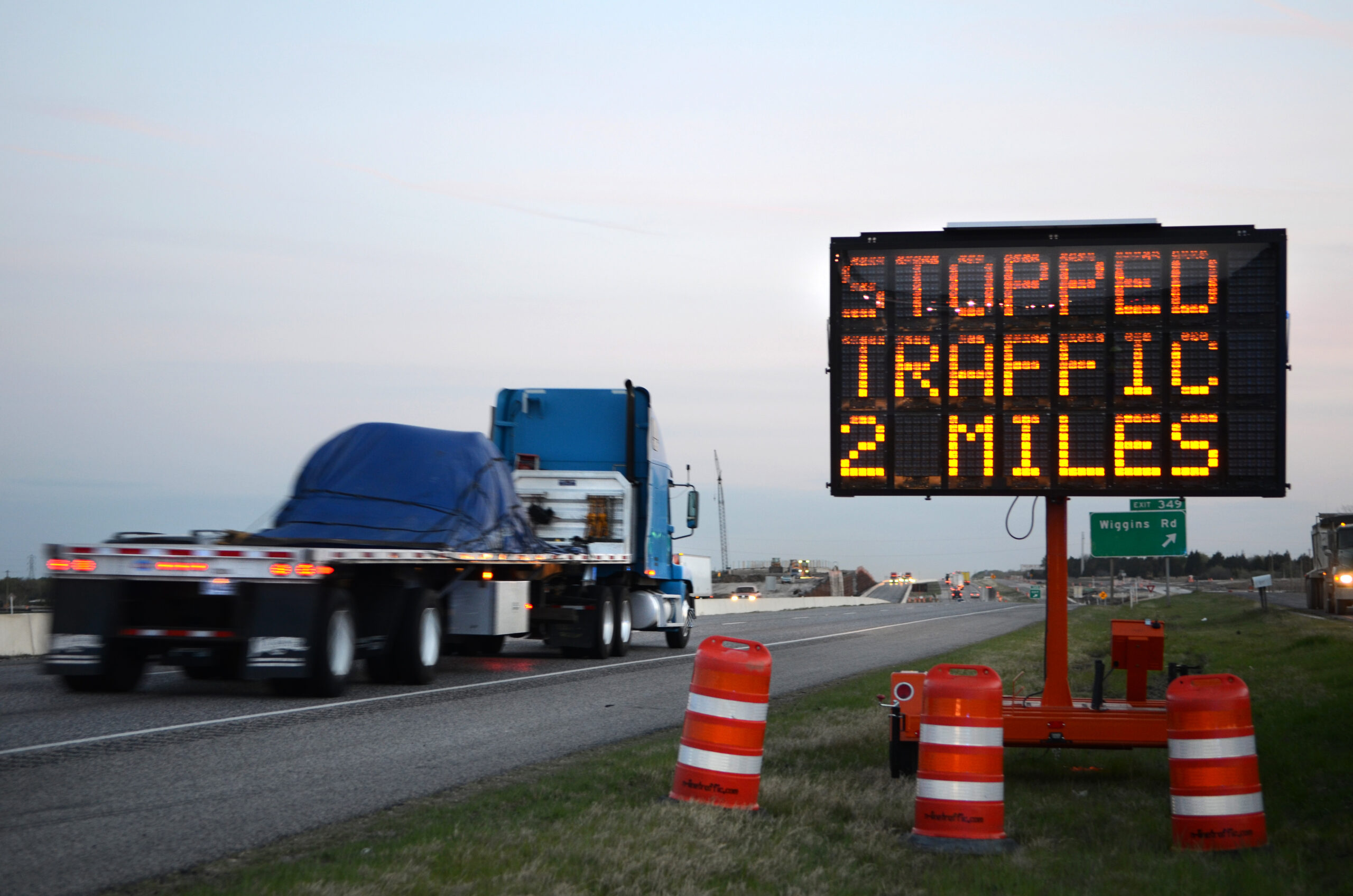 Electronic sign on roadside reads STOPPED TRAFFIC 2 MILES with a semi-truck passing by and orange traffic barrels positioned nearby.