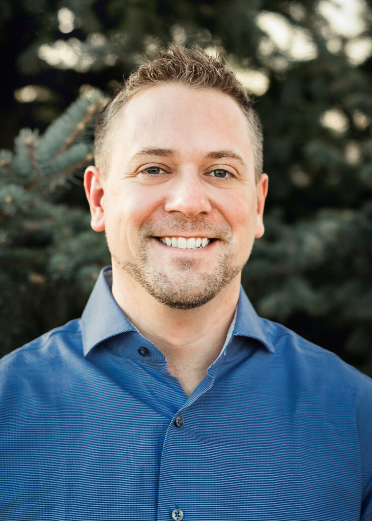A man with short brown hair and a trimmed beard smiles at the camera, wearing a blue button-up shirt, with greenery in the background.