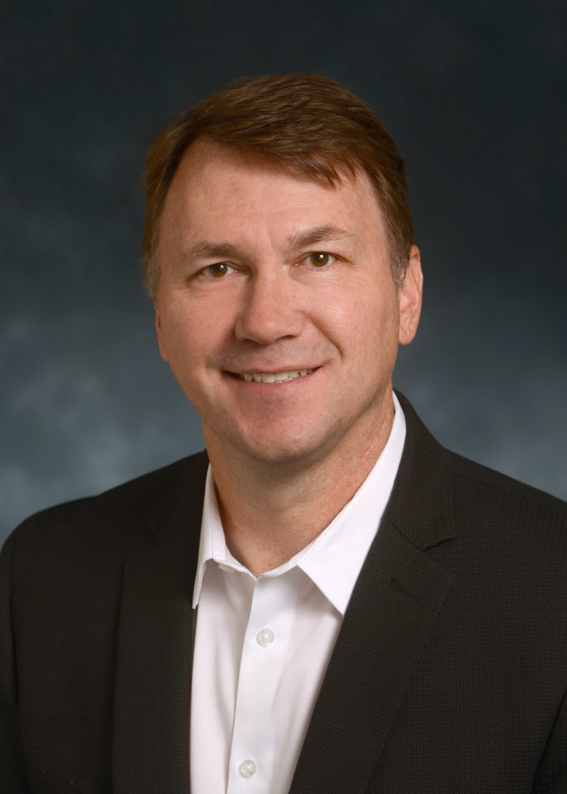A man in a dark suit jacket and white shirt is smiling at the camera against a dark, blurred background.