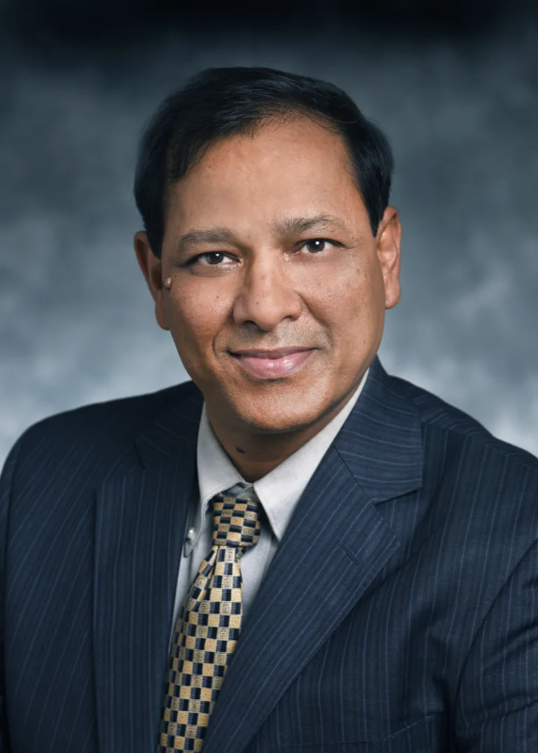 A man in a dark pinstripe suit, light shirt, and patterned tie poses for a professional headshot against a gray background.