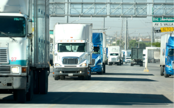 Several semi-trailer trucks drive in both directions on a wide road beneath overhead signs, with more trucks visible in the background.