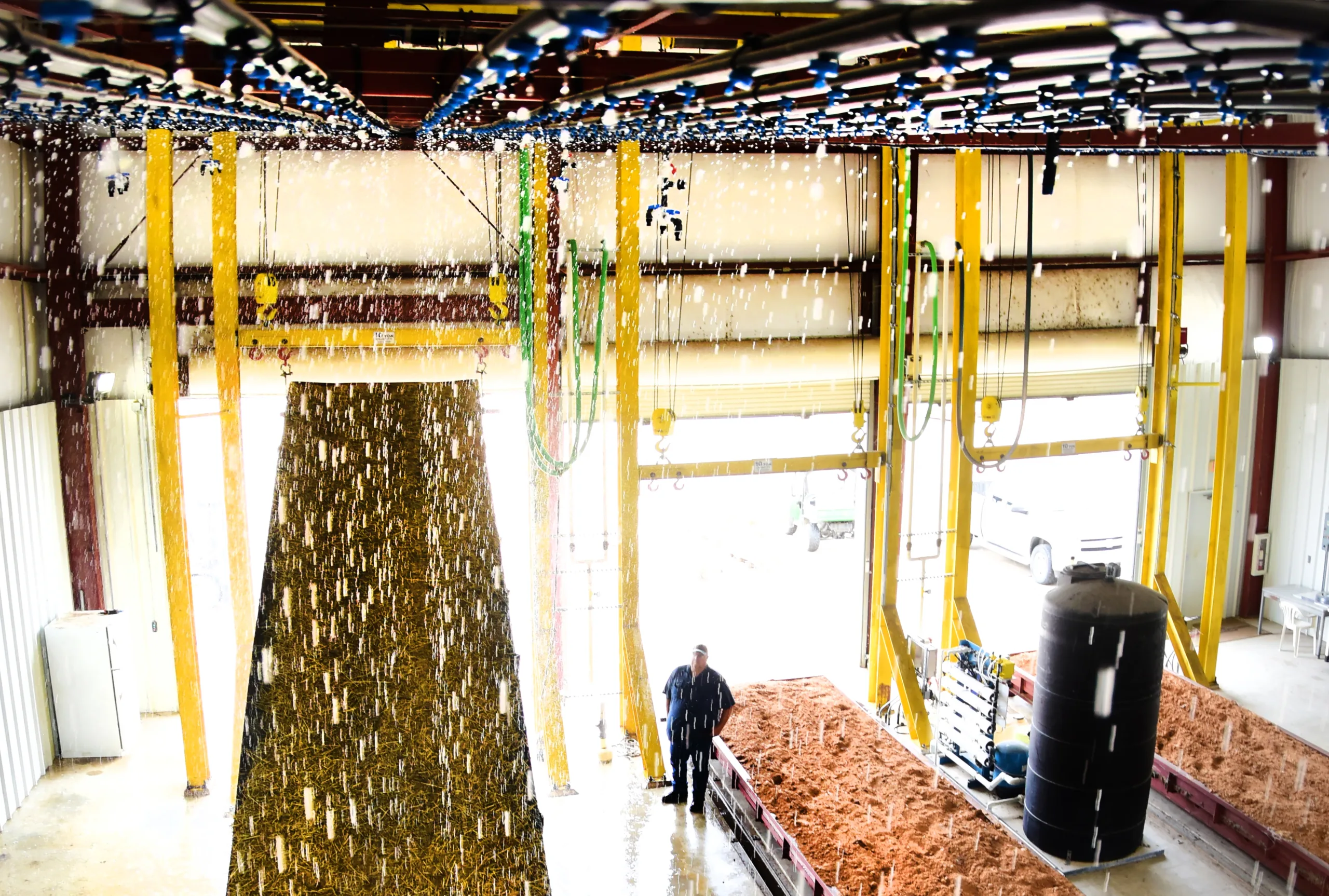 A person stands in a large industrial facility beneath cascading streams of water, with machinery, hoses, and sand beds visible in the background.