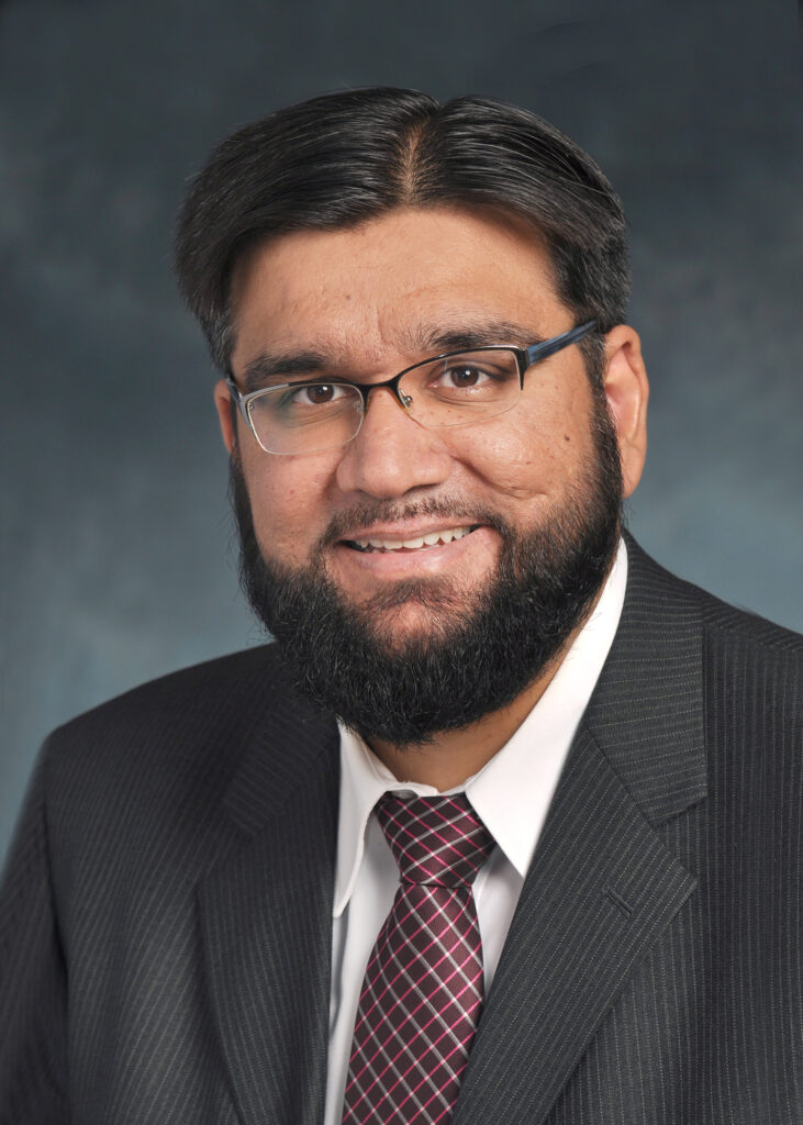 A man wearing glasses, a dark suit, a white shirt, and a patterned tie poses for a professional headshot against a neutral background.