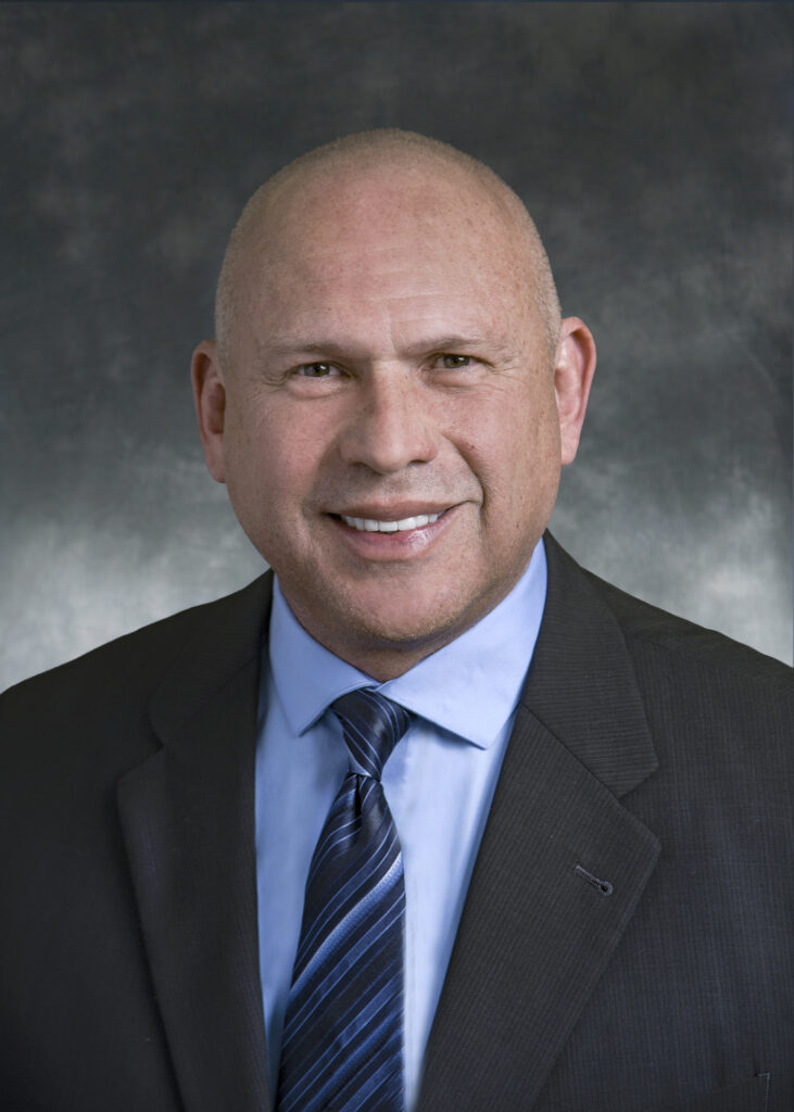 A bald man in a dark suit, light blue shirt, and striped tie smiles at the camera against a dark, mottled background.