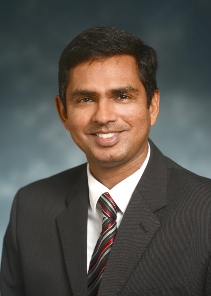 A man in a dark suit, white shirt, and striped tie smiles at the camera against a blurred blue-gray background.