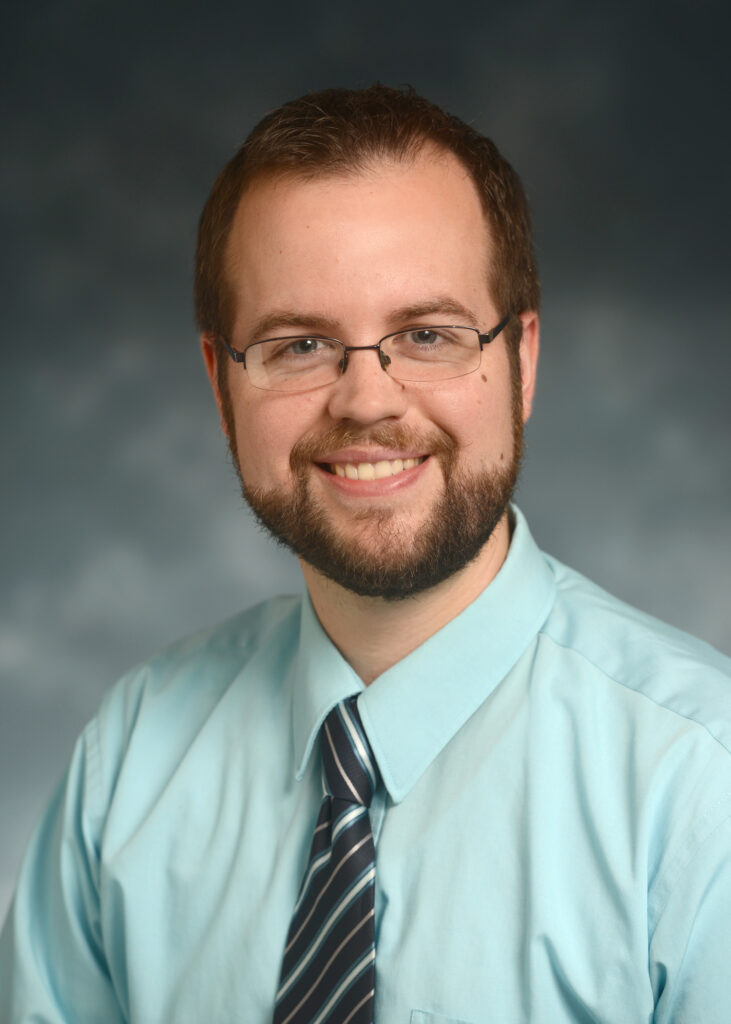 A man with short brown hair, glasses, and a beard, wearing a light blue shirt and striped tie, smiles at the camera against a neutral blurred background.