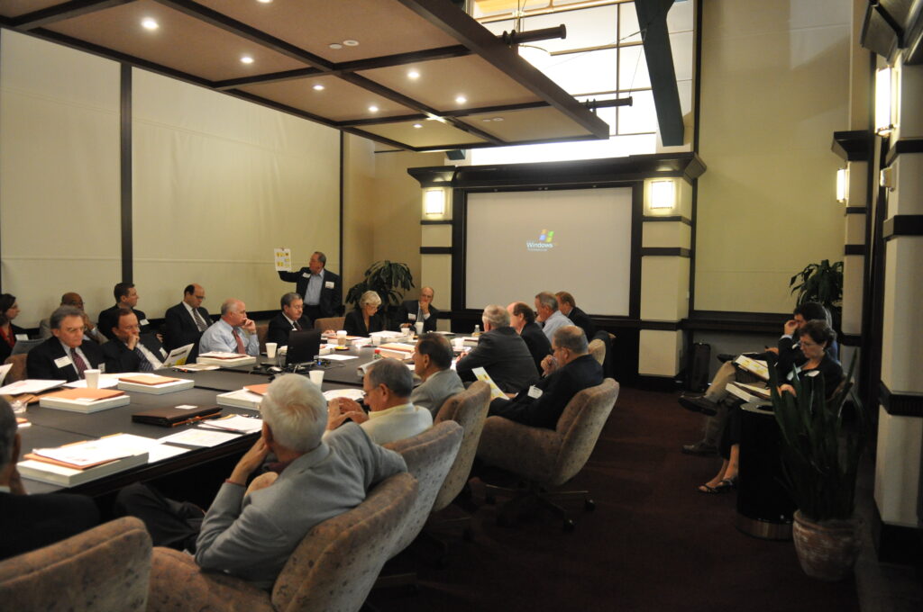 A group of people in business attire sit around a large conference table in a meeting room, with a projector displaying a Windows logo on the screen.