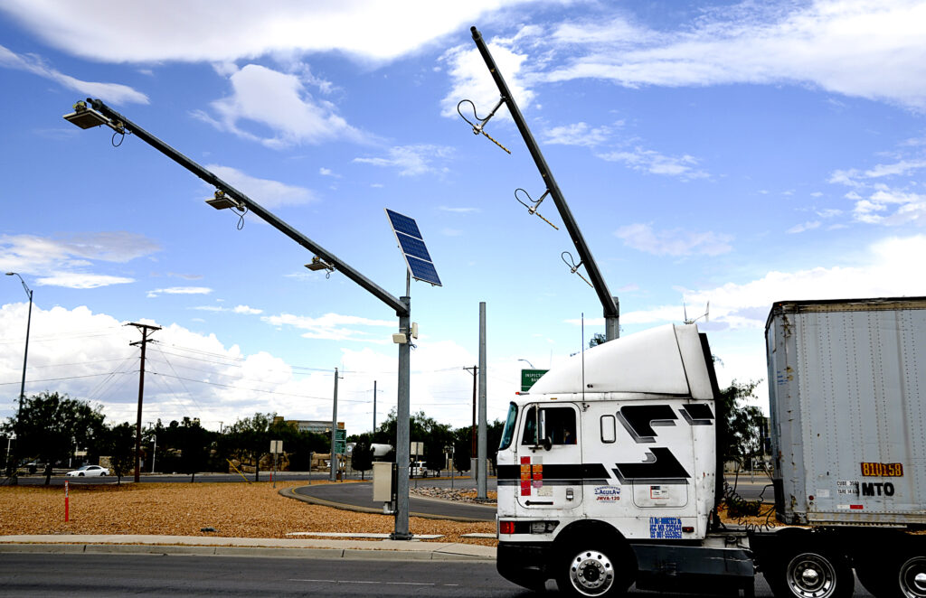 A white semi-truck drives past roadside poles equipped with cameras and solar panels under a partly cloudy sky.