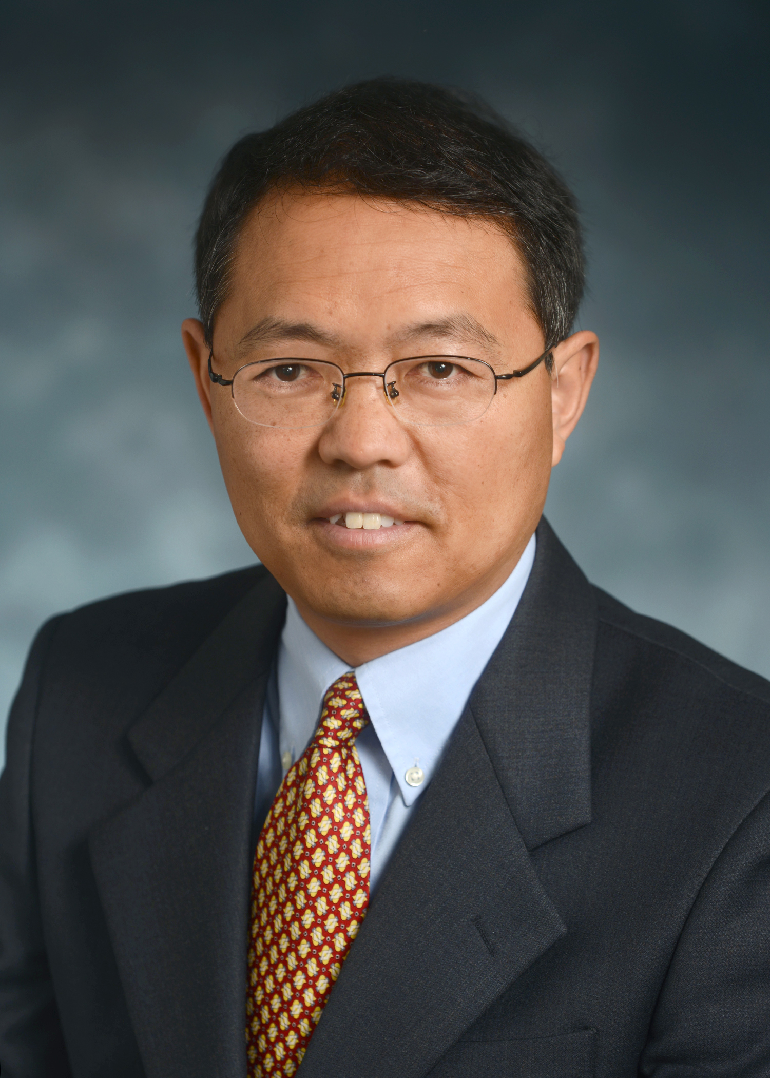A man wearing glasses and a dark suit with a patterned tie poses for a professional headshot against a neutral background.