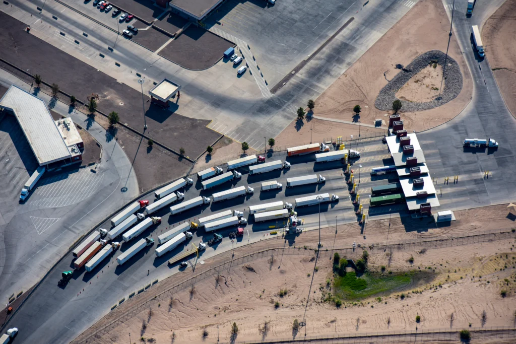 Aerial view of multiple semi-trucks lined up at a checkpoint with several inspection lanes and buildings in a desert area.
