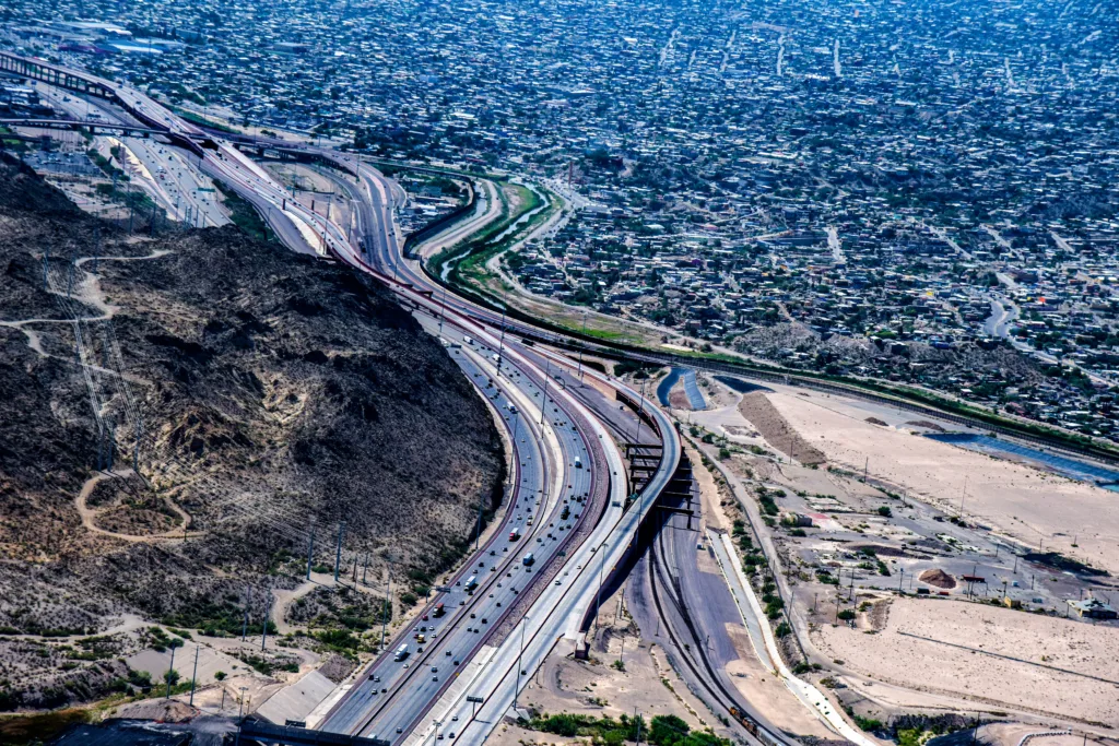 Aerial view of a highway curving around a rocky hill, with a sprawling urban area in the background and a dry, sandy landscape in the foreground.