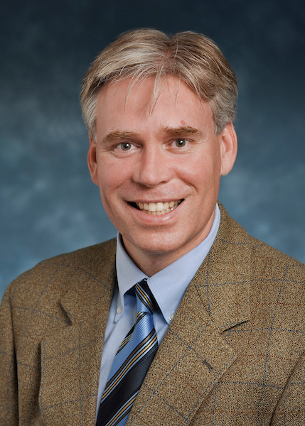 A man with light hair wearing a brown plaid suit jacket, blue shirt, and striped tie, smiles at the camera against a blue background.