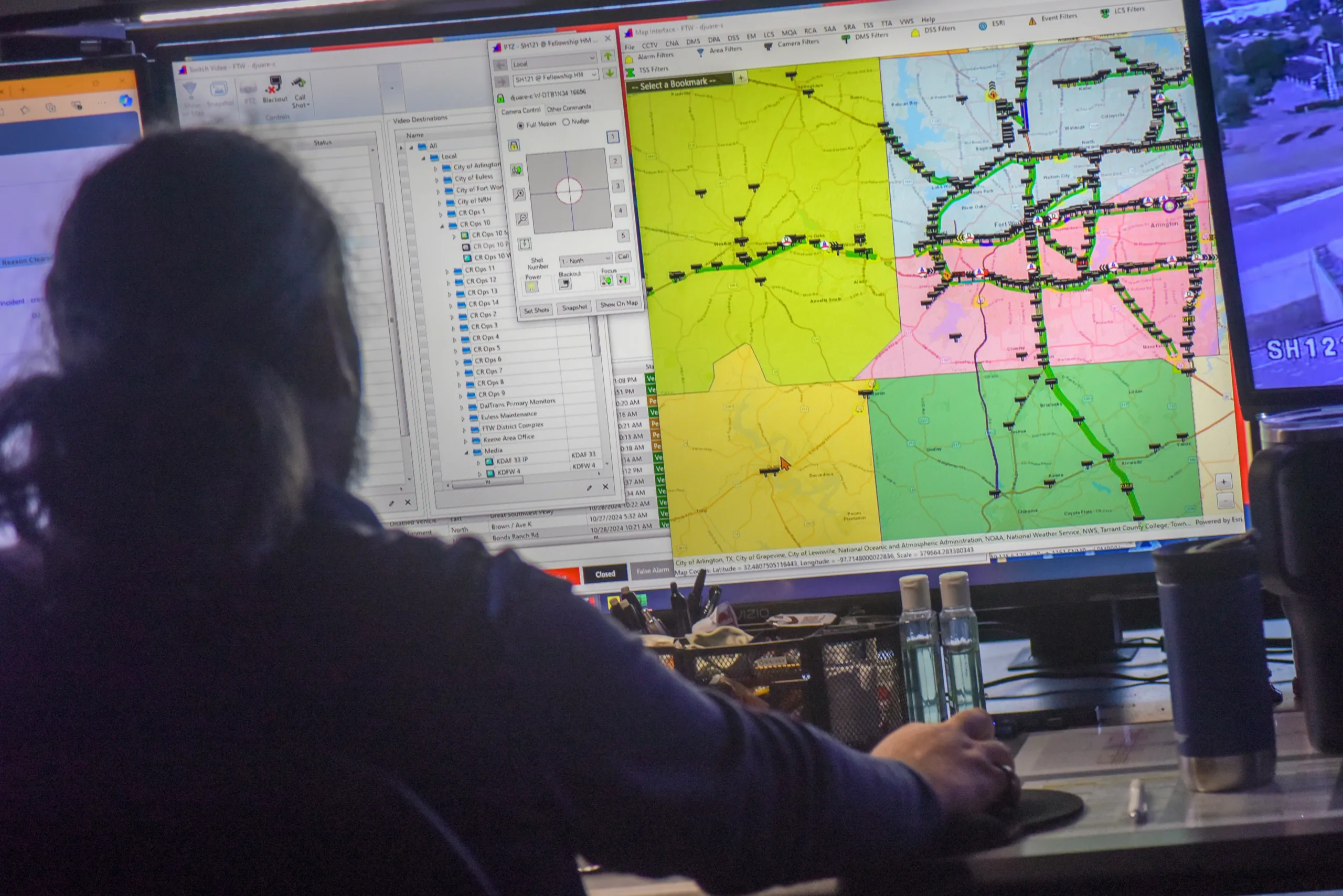 A person sits at a desk monitoring computer screens displaying a color-coded map, data lists, and a live camera feed.