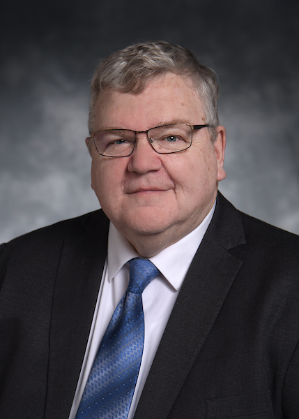A man with gray hair and glasses wearing a dark suit, white shirt, and blue tie poses in front of a gray background.