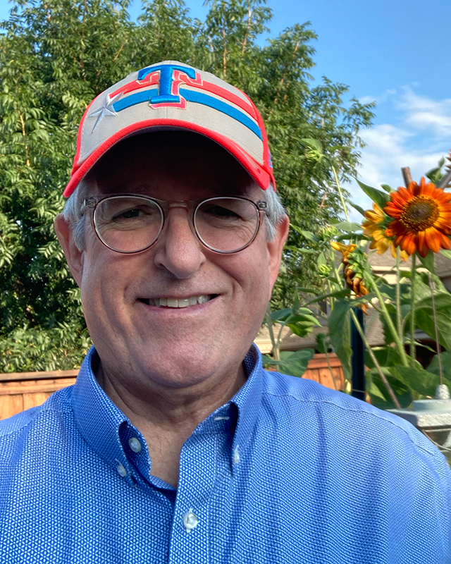 Outdoors of picture Allan Rutter with a large orange flower, green trees, and a blue sky in the background