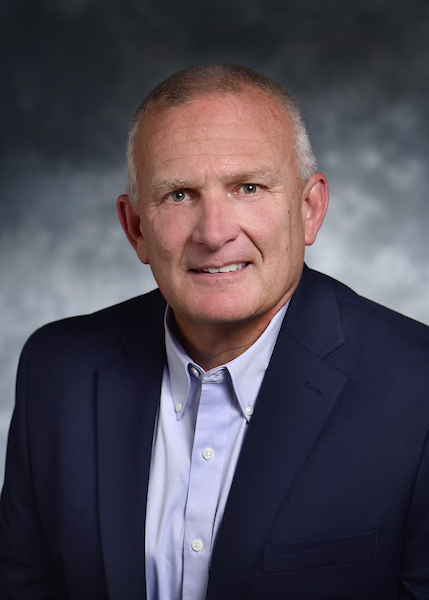 A middle-aged man with short gray hair wearing a navy blazer and light blue shirt, posing against a neutral, blurred background.