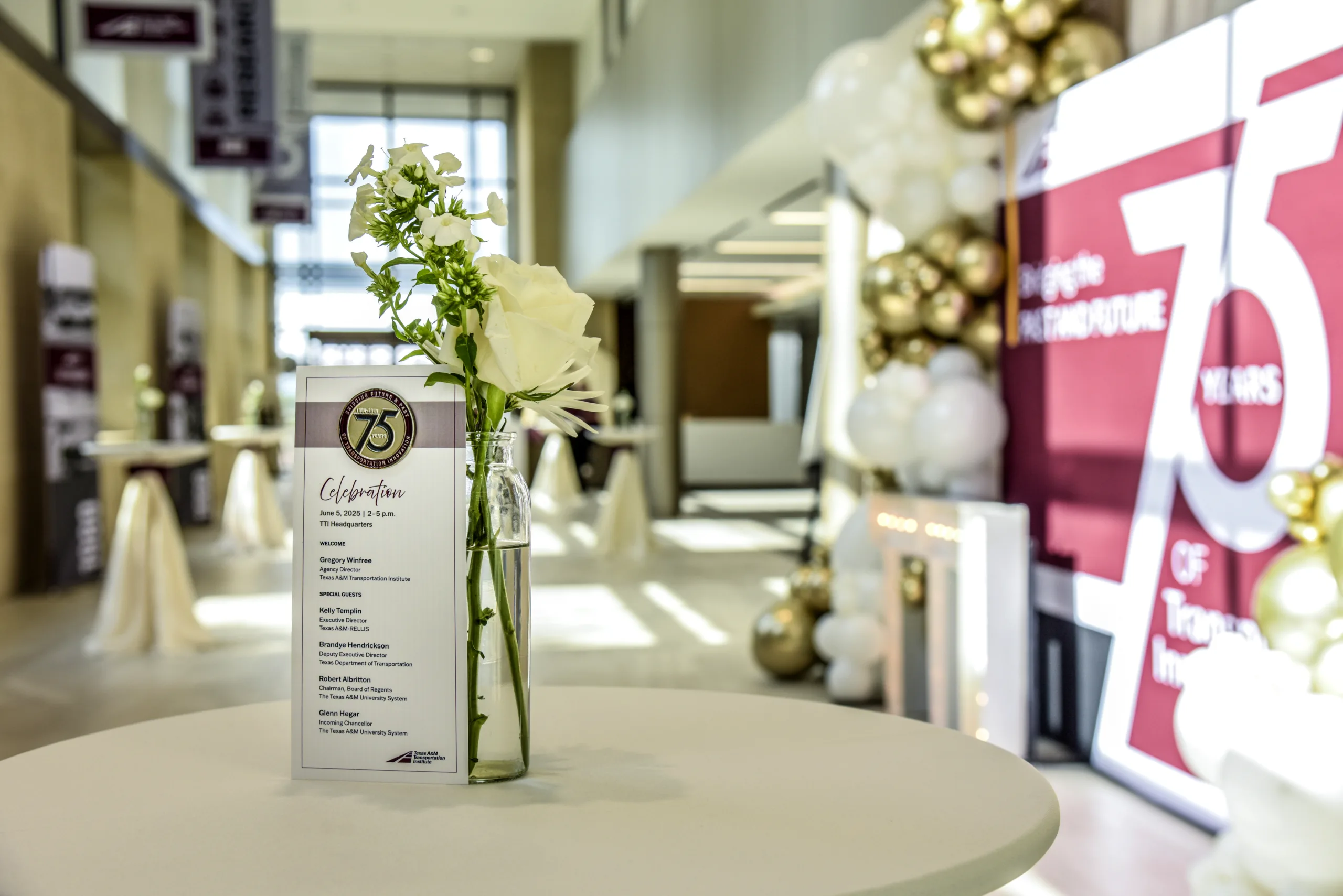 A white rose in a glass vase on a round table with a 75th anniversary celebration sign in a decorated hall.