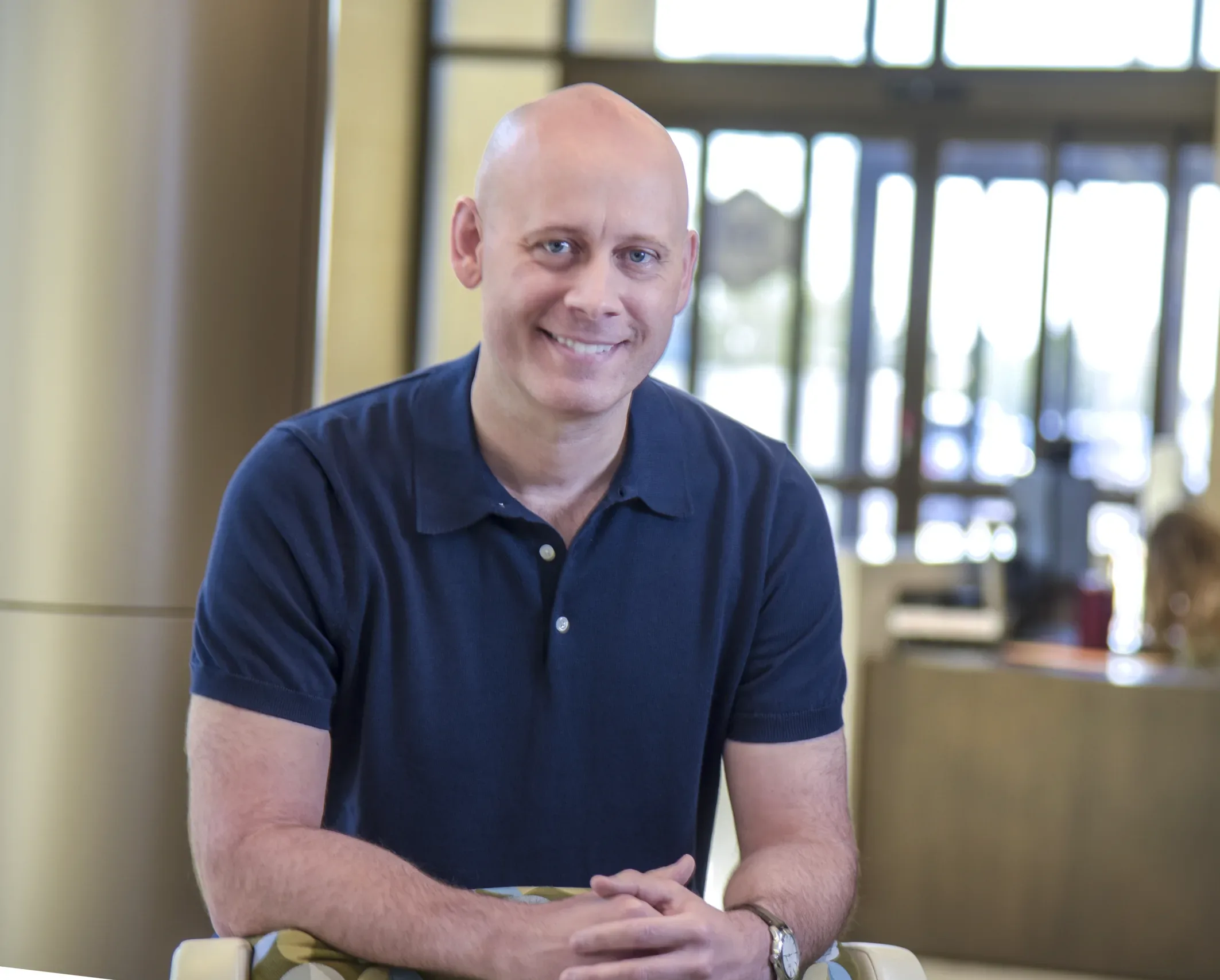 Smiling bald man in a navy polo shirt sits indoors with a blurred office background.