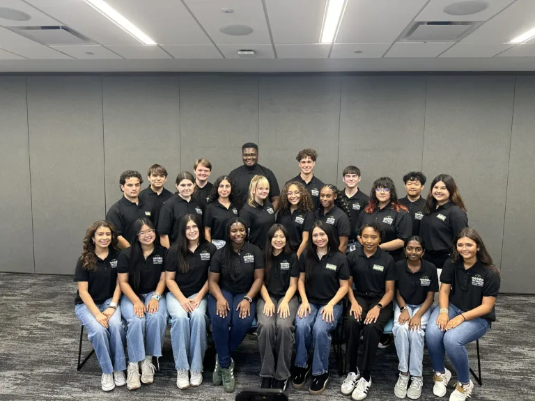 A group of 23 students in matching black shirts pose together indoors, smiling at the camera.