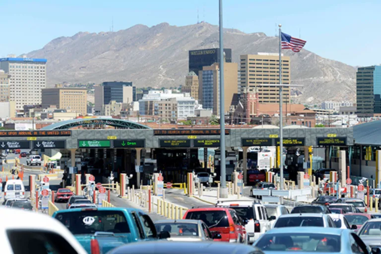 Cars line up at a busy border crossing checkpoint with city buildings and mountains in the background.