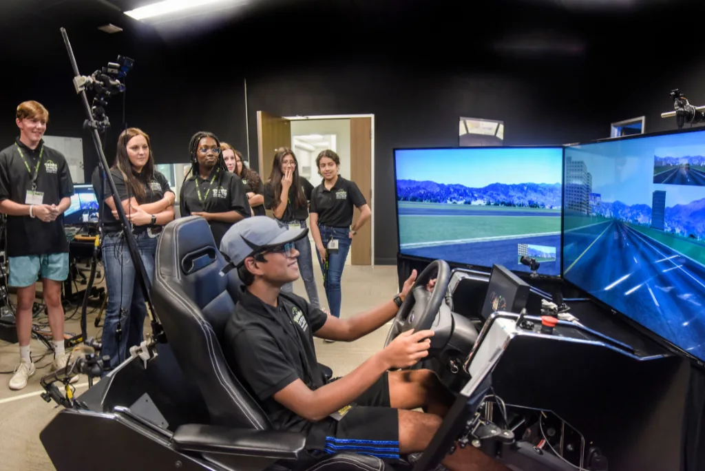 A group of students watch as a person uses a driving simulator with three large monitors in a lab, participating in a youth transportation safety program in Texas.
